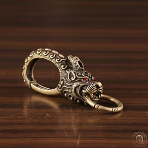 Op de afbeelding: Een messingkleurige metalen sleutelhanger met een gebeeldhouwde leeuwenkop met gedetailleerde wervelpatronen en twee kleine, ingelegde rode stenen als ogen. De bek van de leeuw houdt een ronde ring vast aan de voorzijde.