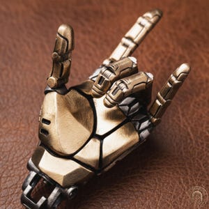 Peut inclure: Une figurine de main robotique aux tons or et argent métallisés, posée dans un geste de rock and roll. La main mécanique présente des doigts segmentés, des articulations visibles et une finition en métal brossé avec des lignes sombres.