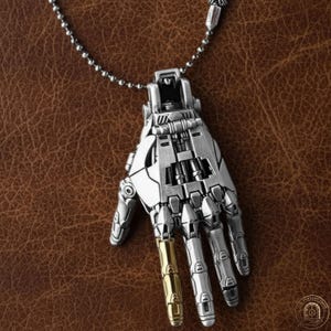 Może przedstawiać: Srebrny metalowy wisiorek w kształcie robota. Wisiorek ma skomplikowane detale mechaniczne, z jednym złotym palcem. Naszyjnik wisi na srebrnym łańcuszku kulkowym, na tle brązowej skóry.