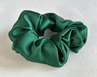 May include: A dark emerald green scrunchie made of a silky fabric. The hair accessory is circular and has a gathered, ruffled appearance. The scrunchie is a solid color and appears to be made of satin.
