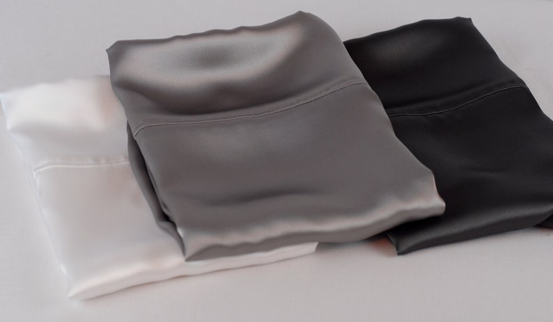 May include: Three satin pillowcases in white, gray, and black. The pillowcases are folded and stacked on top of each other.