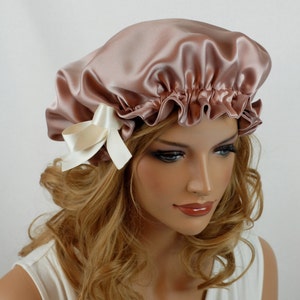 May include: A blush pink satin sleep cap with a white satin bow. The cap is designed to protect hair while sleeping.