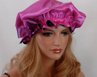 Op de afbeelding: Een felroze satijnen bonnet met een gerimpelde rand en een contrasterende paarse bies. De bonnet is ontworpen om het haar te beschermen en wordt getoond op een mannequin met blond haar.