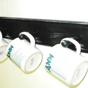 May include: Black wooden mug rack with three white mugs hanging from hooks. The mugs have the names "John", "Ann", and "Nicole" written on them.