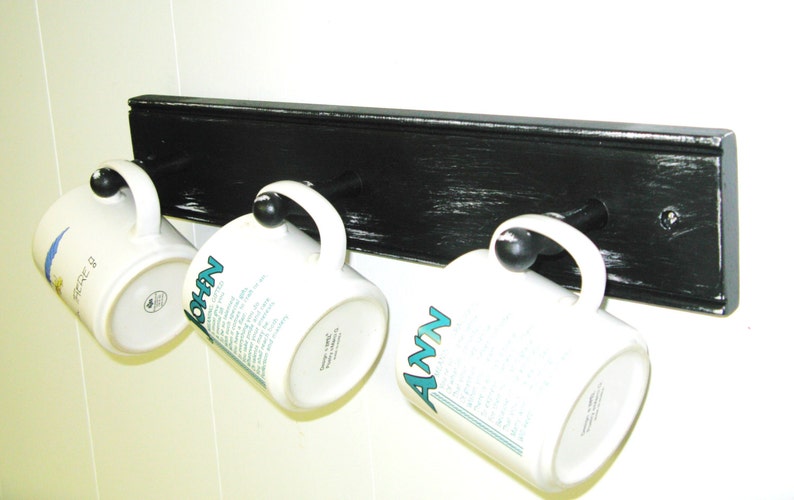 May include: Black wooden mug rack with three white mugs hanging from hooks. The mugs have the names "John", "Ann", and "Nicole" written on them.