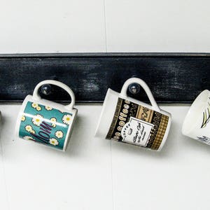 May include: Four ceramic mugs hang on a black wooden rack. The mugs have different designs, including a blue mug with a snowflake pattern, a green mug with a floral pattern, a white mug with a coffee design, and a white mug with a woman in a dress design.