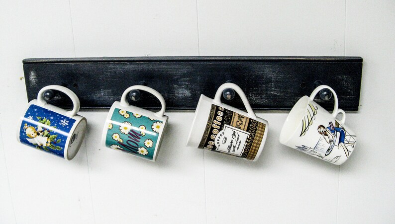 May include: Four ceramic mugs hang on a black wooden rack. The mugs have different designs, including a blue mug with a snowflake pattern, a green mug with a floral pattern, a white mug with a coffee design, and a white mug with a woman in a dress design.