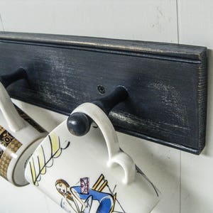 May include: Two ceramic mugs hang on a black wooden rack with metal hooks. The mugs have different designs. One mug has a black and gold design with a floral pattern. The other mug has a white background with a colorful illustration of a woman.