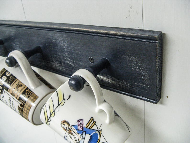 May include: Two ceramic mugs hang on a black wooden rack with metal hooks. The mugs have different designs. One mug has a black and gold design with a floral pattern. The other mug has a white background with a colorful illustration of a woman.