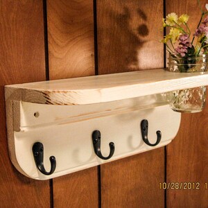 May include: White wooden shelf with three hooks and a small glass jar for holding flowers. The shelf is mounted on a wood panel wall.