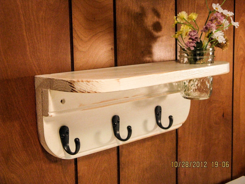 May include: White wooden shelf with three hooks and a small glass jar for holding flowers. The shelf is mounted on a wood panel wall.