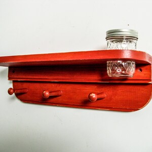 May include: Red wooden shelf with three hooks and a small glass jar on the top shelf.