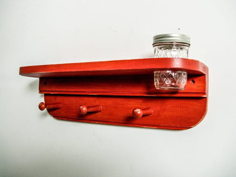 May include: Red wooden shelf with three hooks and a small glass jar on the top shelf.