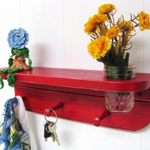 May include: Red wooden wall shelf with two hooks, a small blue flower figurine, and a mason jar vase with yellow flowers.