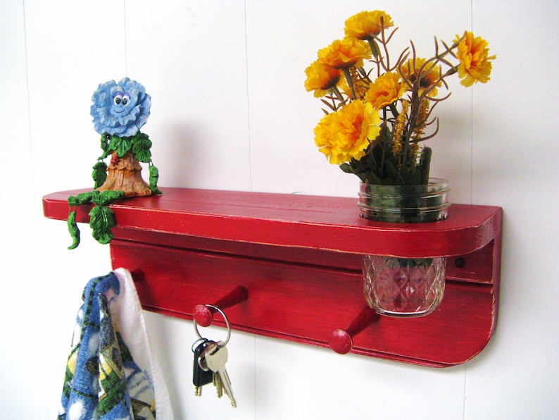 May include: Red wooden wall shelf with two hooks, a small blue flower figurine, and a mason jar vase with yellow flowers.