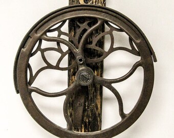 May include: A vintage cast iron wheel with a decorative, intricate design. The wheel is mounted on a wooden block and has a metal axle.