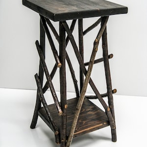 May include: A rustic, dark brown wooden plant stand. The stand features a square top and a lower shelf, both made of solid wood. The legs and supports are crafted from natural branches, creating a unique, handcrafted look.