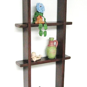 May include: A dark brown wooden wall shelf with four shelves. The shelves are decorated with various small figurines, including a blue flower, a green and pink bottle, a small angel, and a black dog.