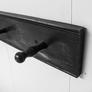 May include: A black wooden coat rack with three hooks. The rack is mounted on a white wall.