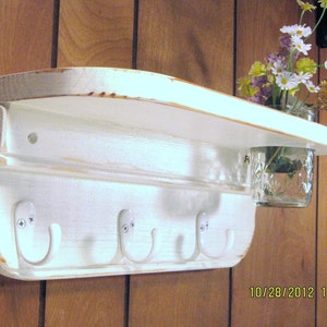 May include: White wooden shelf with four hooks and a glass jar for flowers. The shelf has a distressed white finish and is mounted on a wood wall.