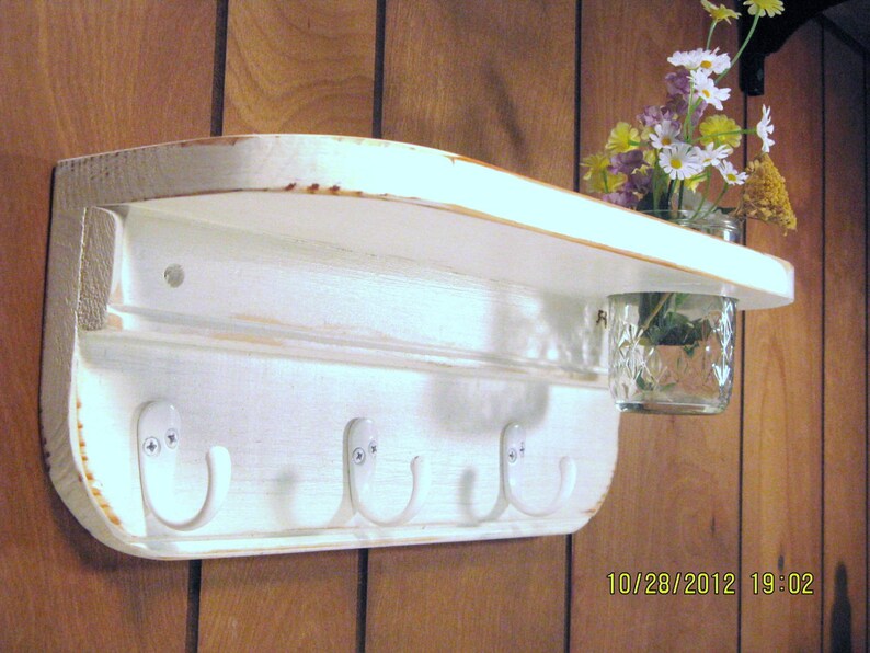 May include: White wooden shelf with four hooks and a glass jar for flowers. The shelf has a distressed white finish and is mounted on a wood wall.