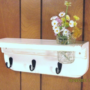 May include: White wooden shelf with three hooks and a glass jar for storage. The shelf is mounted on a wood panel wall.  The shelf has a distressed white paint finish.  The hooks are black metal.  The jar is clear glass with a diamond pattern.  The shelf is decorated with a bouquet of white and yellow flowers.