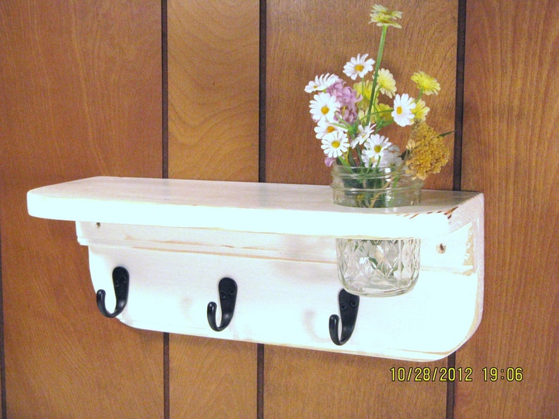May include: White wooden shelf with three hooks and a glass jar for storage. The shelf is mounted on a wood panel wall.  The shelf has a distressed white paint finish.  The hooks are black metal.  The jar is clear glass with a diamond pattern.  The shelf is decorated with a bouquet of white and yellow flowers.