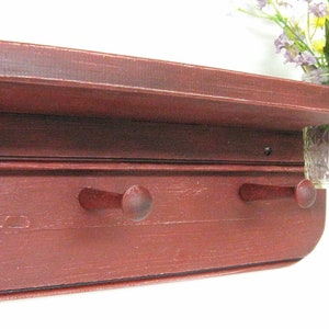 May include: A red wooden shelf with two hooks and a glass jar attached to the side. The shelf has a distressed finish and is perfect for adding rustic charm to any room.