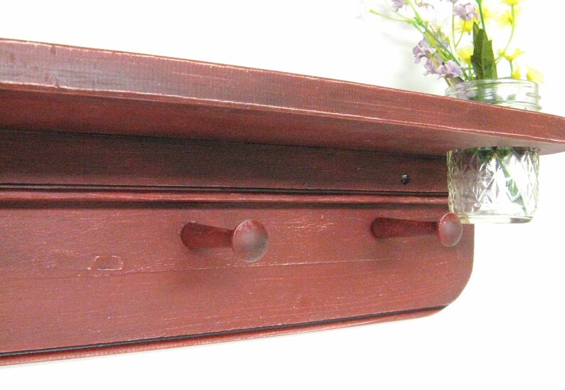 May include: A red wooden shelf with two hooks and a glass jar attached to the side. The shelf has a distressed finish and is perfect for adding rustic charm to any room.