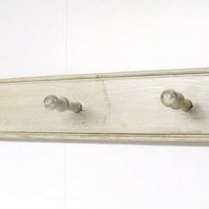 May include: White wooden wall mounted coat rack with three knobs. The rack has a distressed finish and the knobs are painted a light gray.