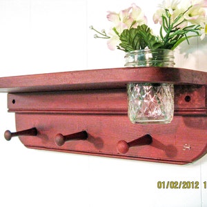 May include: A red wooden shelf with three hooks and a glass jar for a vase. The shelf has a distressed finish and is mounted on a white wall. The jar is filled with white flowers.
