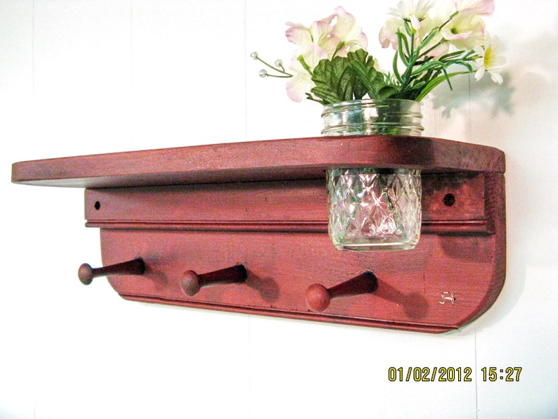 May include: A red wooden shelf with three hooks and a glass jar for a vase. The shelf has a distressed finish and is mounted on a white wall. The jar is filled with white flowers.