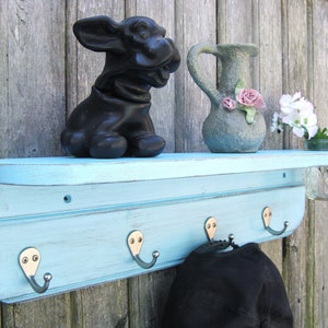 May include: A light blue wooden shelf with a curved top and four hooks. The shelf has a distressed finish and is mounted on a wood wall. There is a black ceramic dog figurine, a gray ceramic pitcher with pink flowers, and a glass jar with white flowers on the shelf.