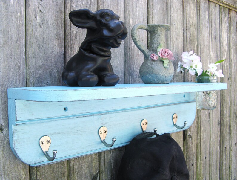 May include: A light blue wooden shelf with a curved top and four hooks. The shelf has a distressed finish and is mounted on a wood wall. There is a black ceramic dog figurine, a gray ceramic pitcher with pink flowers, and a glass jar with white flowers on the shelf.