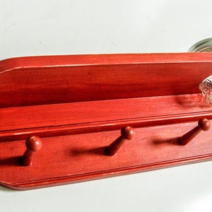 May include: Red wooden wall shelf with three hooks and a small glass jar on top. The shelf is painted a bright red color and has a slightly curved top.