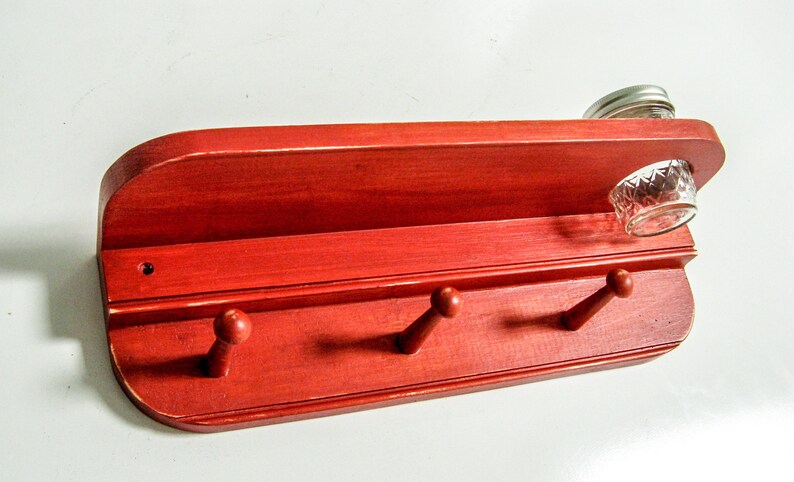 May include: Red wooden wall shelf with three hooks and a small glass jar on top. The shelf is painted a bright red color and has a slightly curved top.