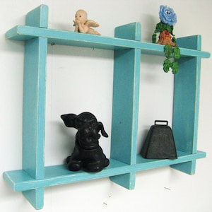 May include: A light blue wooden shelf unit with three shelves. The shelves are divided into two sections by a vertical divider. There are various decorative items on the shelves, including a small angel figurine, a flower figurine, a black dog figurine, and a metal bell.