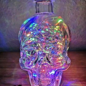 May include: A clear glass bottle shaped like a skull, with a cork stopper, illuminated by multi-coloured LED fairy lights. The lights inside the bottle create a vibrant display of colour. The bottle is placed on a wooden surface.