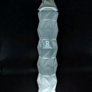 May include: A tall, geometric-patterned bottle with a light-up base. The bottle is a pale grey colour and features a repeating diamond design. The letter "B" is visible on the front. The top of the bottle has a unique, angular design.