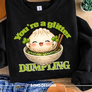 Puede incluir: Sudadera negra con un diseño de una bola de masa sonriente en un cuenco con palillos. El texto "You're a glitter dumpling" y "DUMPLING" son verdes. Un diseño divertido con temática de comida.