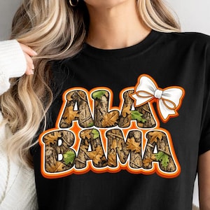 Pode incluir: Camiseta preta com a palavra "ALABAMA" em um padrão de camuflagem com uma borda laranja. Uma fita branca está colocada acima da palavra. A camiseta é usada por uma pessoa com cabelo loiro e um cardigã branco.