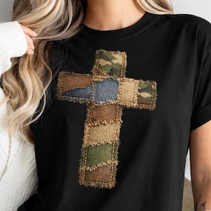 May include: Black t-shirt with a patchwork cross design. The cross is made from various fabrics, including camouflage, denim, and burlap. The shirt has a classic crew neck.