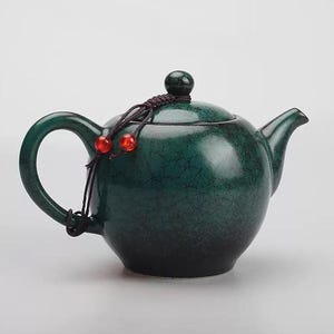 Puede incluir: Una tetera de cerámica verde oscuro con cuerpo redondeado, asa curva y boquilla. La tapa está rematada con un pequeño pomo y un cordón decorativo con dos cuentas rojas. La tetera tiene un acabado craquelado.