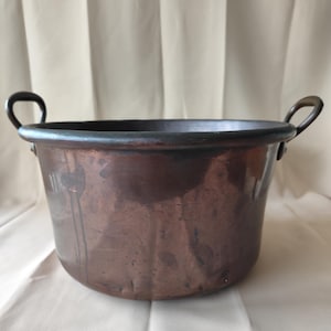 Puede incluir: Un caldero de cobre con dos asas curvas. La olla tiene un cuerpo ancho y redondeado y un borde oscuro. El cobre tiene un color marrón rojizo intenso con algunas zonas más oscuras, lo que sugiere edad o uso. El caldero es probablemente para cocinar o decorar.
