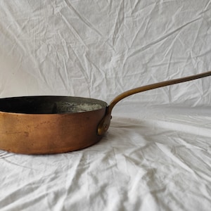 Op de afbeelding: Een koperen steelpan met een lange, gebogen handgreep. De pan heeft een brede, ondiepe kom en een donker interieur. De handgreep is aan de zijkant van de pan bevestigd en buigt omhoog. De pan staat tegen een witte achtergrond.