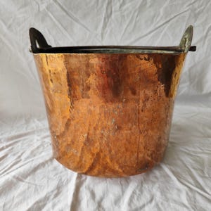 Puede incluir: Un cubo de cobre con dos asas curvas. El cubo tiene un cuerpo ancho y redondeado y un borde oscuro. El cobre tiene un color marrón rojizo cálido con variaciones de tono, lo que sugiere edad y uso. Las asas son de un metal más oscuro.