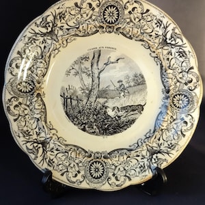 Può includere: Piatto decorativo antico con una scena di caccia in bianco e nero al centro, incorniciato da un bordo floreale e fogliaceo dettagliato. Il piatto ha un bordo smerlato e il testo "CHASSE AUX PERDRIX" è visibile.