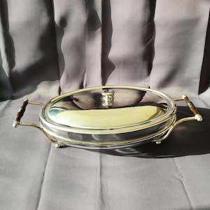 Może przedstawiać: Owalna srebrna patera do serwowania z szklaną pokrywą. Naczynie ma dwa uchwyty z ciemnobrązowymi drewnianymi akcentami. Pokrywa ma mały srebrny uchwyt. Naczynie stoi na szarej powierzchni.