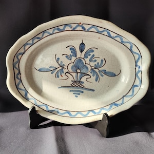 May include: Oval ceramic serving dish with a white base and blue floral design. The rim features a wavy blue pattern and a brown border. The dish is supported by a black stand, showcasing its decorative and functional aspects.