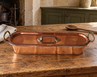 May include: An elongated rectangular copper roasting pan with a matching lid. The pan features two curved brass side handles and a central brass handle on the lid. The metal surface shows a warm, polished copper finish with visible rivets.